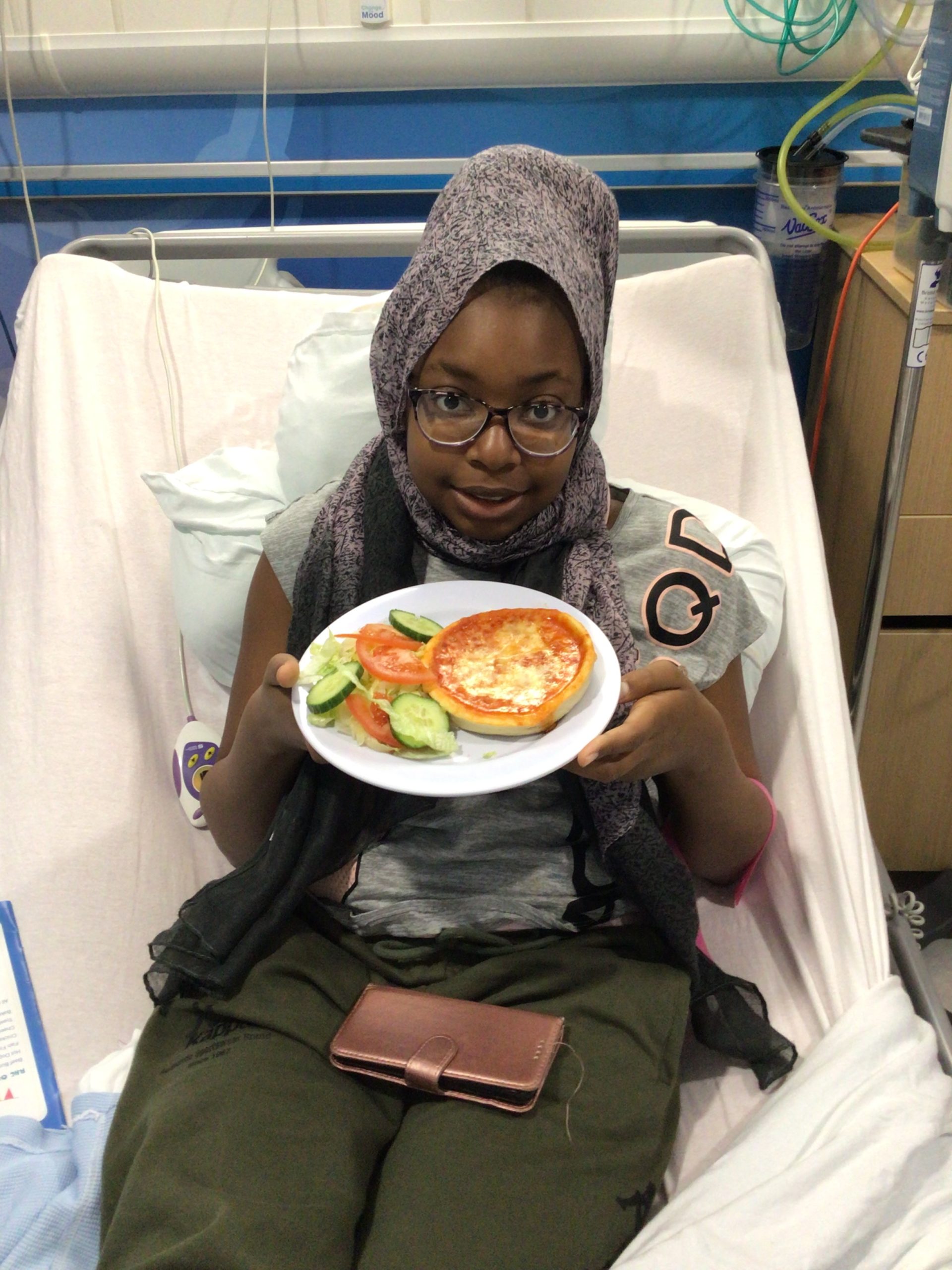 Girl saitting on hospital bed holding plate with pizza and salad