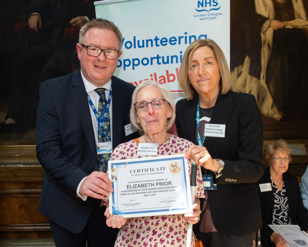 Elizabeth Prior holds her long service award, with NHSGGC Deputy Chief Executive William Edwards and Strategic Lead for Volunteering, Fiona Smith behind her.