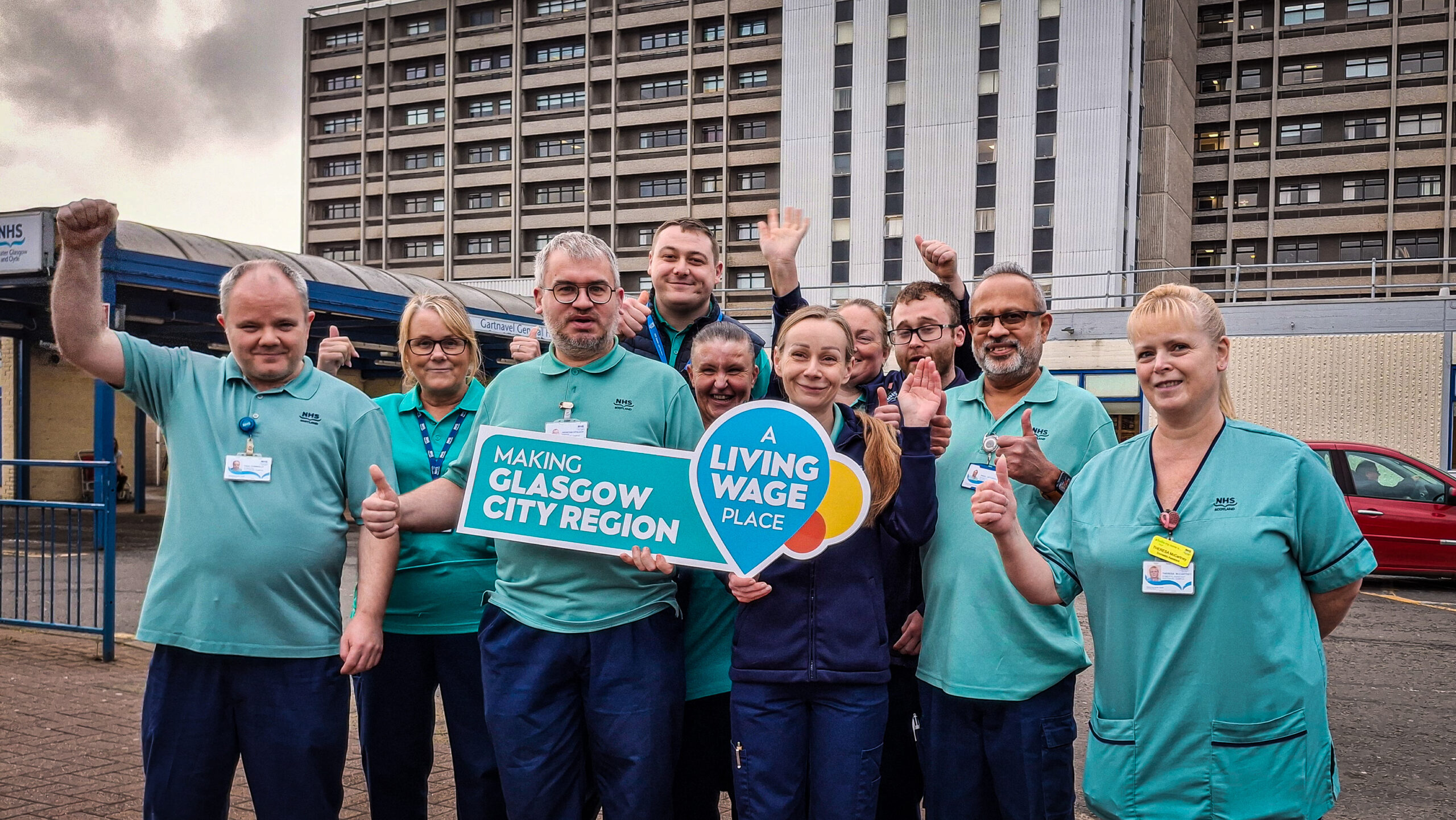 Members of staff standing in their uniforms, smiling and waving outside Gartnavel General Hospital. Two people at the front are holding a sign that reads 'Making Glasgow City Region A Living Wage Place'