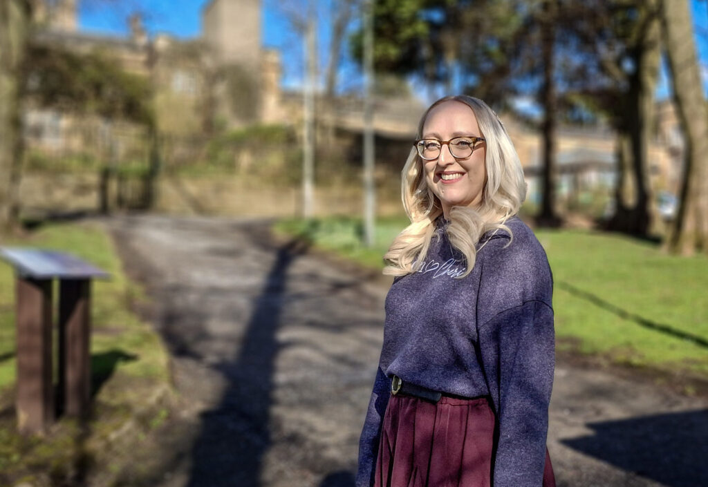 Alison Pugh, dressed in purple sweater and burgundy skirt, stands smiling in the grounds of Gartnavel Royal Hospital, with the sun casting shadows from the trees.