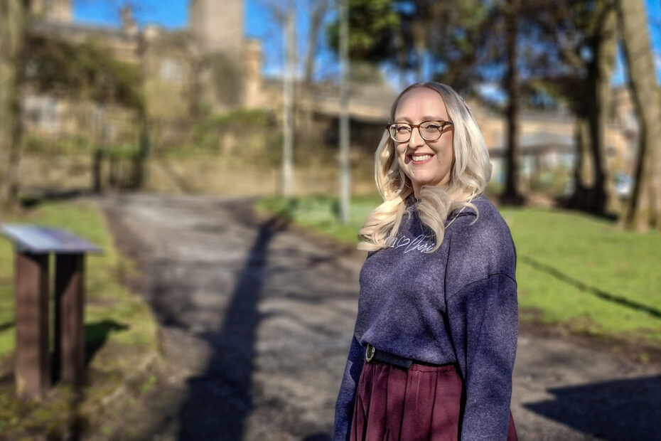 Alison Pugh, dressed in purple sweater and burgundy skirt, stands smiling in the grounds of Gartnavel Royal Hospital, with the sun casting shadows from the trees.