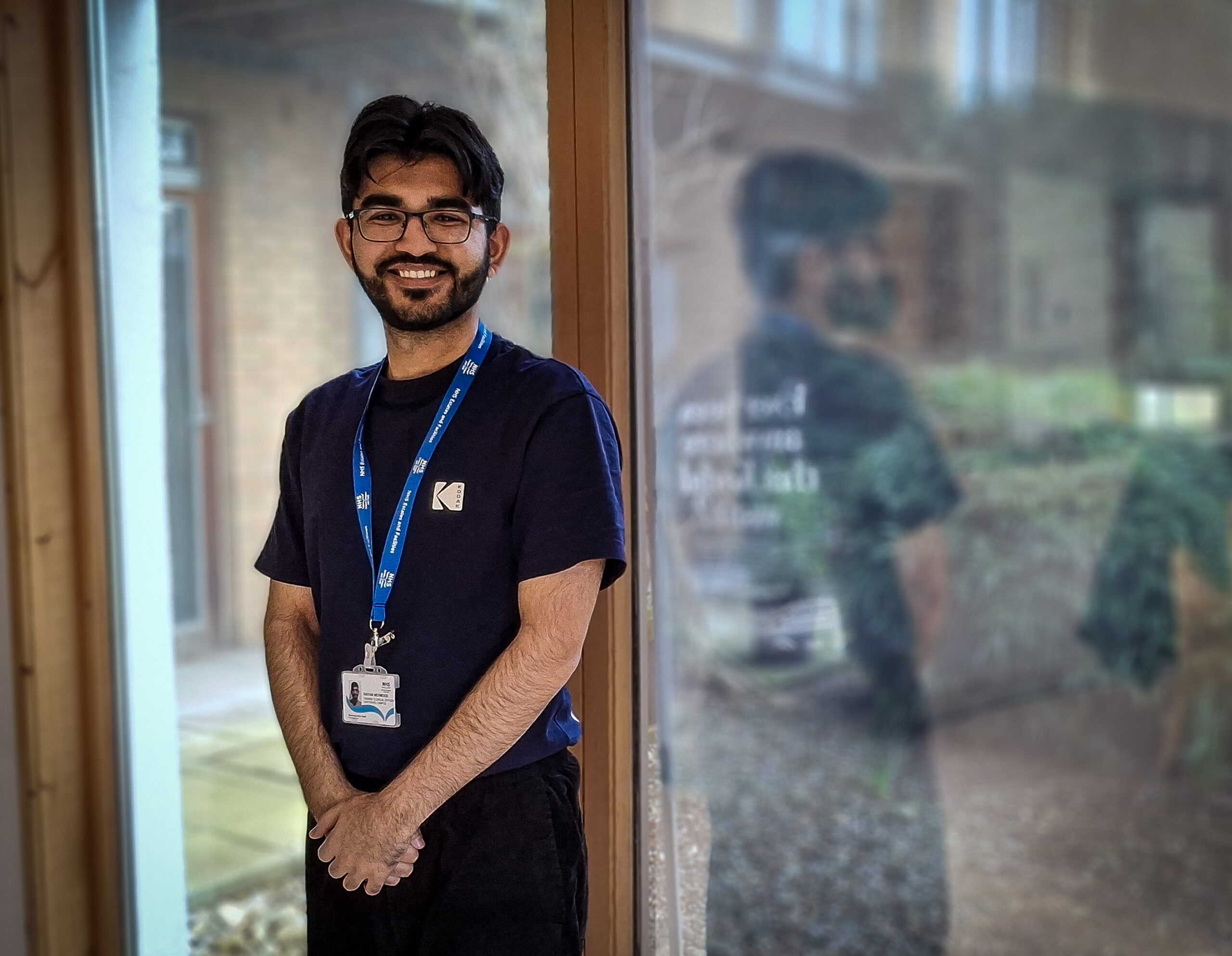 Raiyan Mehmood smiles as he stands next to a large window at the Centre for Integrated Care. The centre's garden can be seen outside.