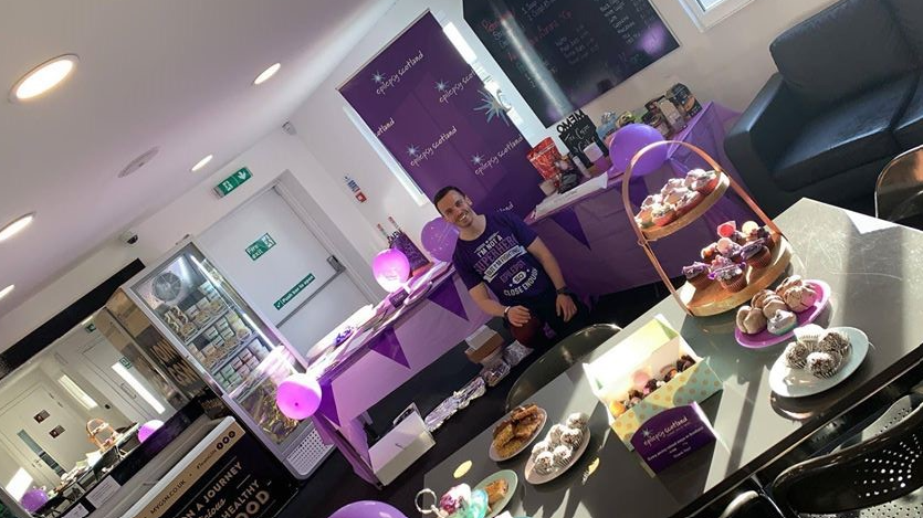A smiling volunteer in a purple T-shirt attends a stall with cakes and snacks at a previous Purple Day celebration. Picture courtesy of Epilepsy Scotland