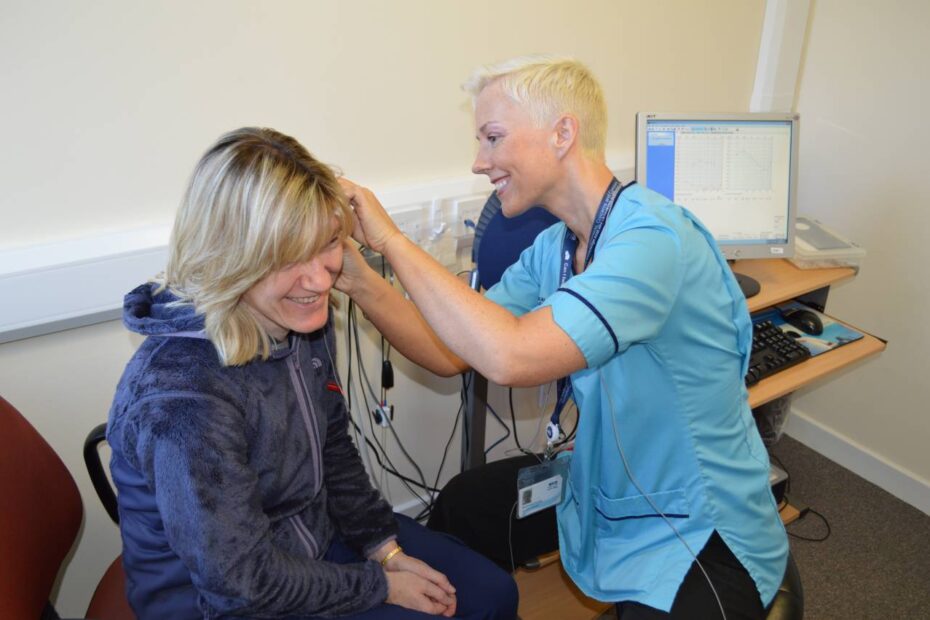 Staff member on right, smiling with short hair, carries out an examination on a patient. during an audiology appointment.