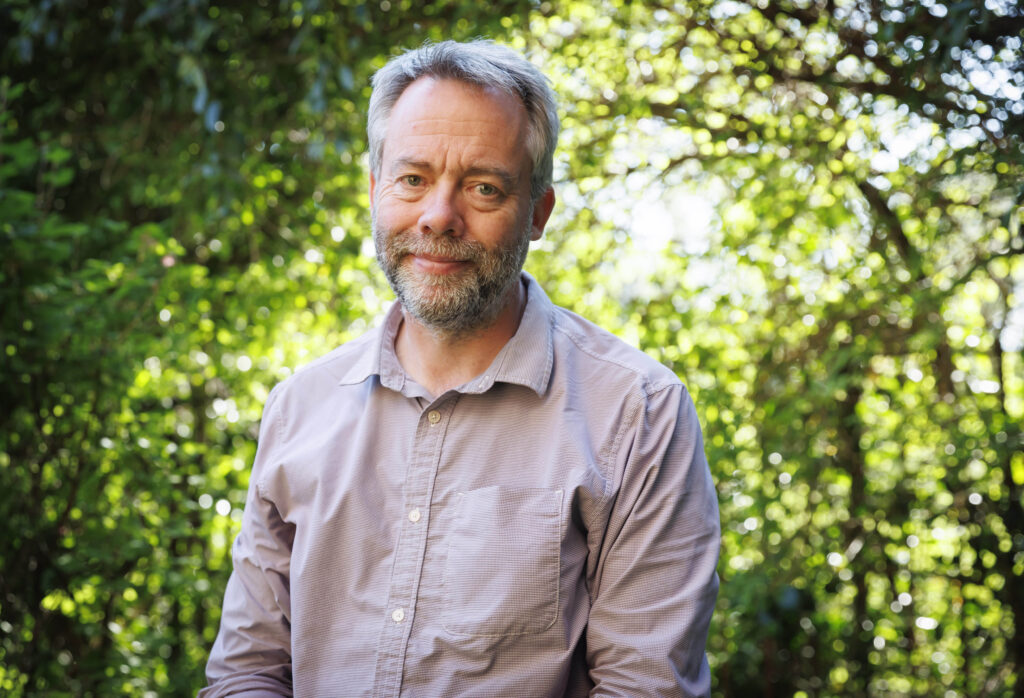 Portrait of Colin Mearns, wearing an open-necked shirt and standing in front of a background of trees and shrubs.