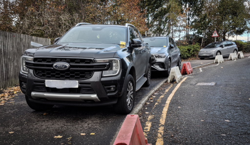 Cars parked on a pavement on hospital grounds. Double yellow lines are visible, and bollards on the road preventing access have been moved so that the cars could park. Two of the cars have parking tickets on the windscreen.