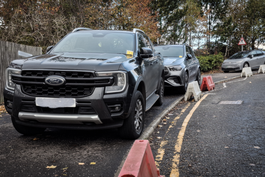 Cars parked on a pavement on hospital grounds. Double yellow lines are visible, and bollards on the road preventing access have been moved so that the cars could park. Two of the cars have parking tickets on the windscreen.