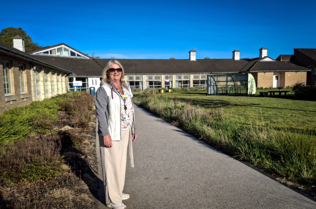 Lesley Donnelly stands on a path between two areas of border that will be left untouched for No Mow May. In the background are ward buildings at Leverndale.