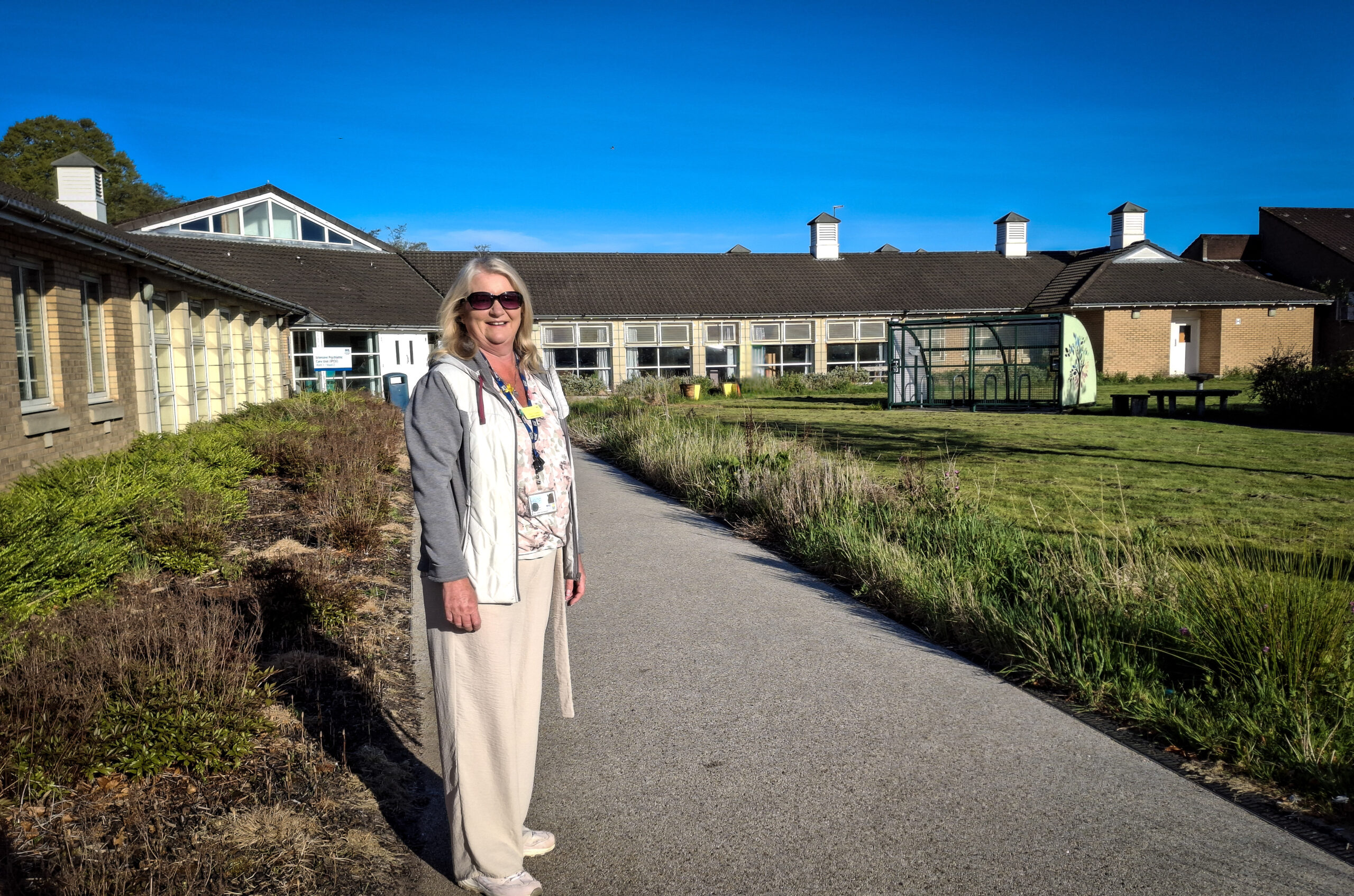 Lesley Donnelly stands on a path between two areas of border that will be left untouched for No Mow May. In the background are ward buildings at Leverndale.