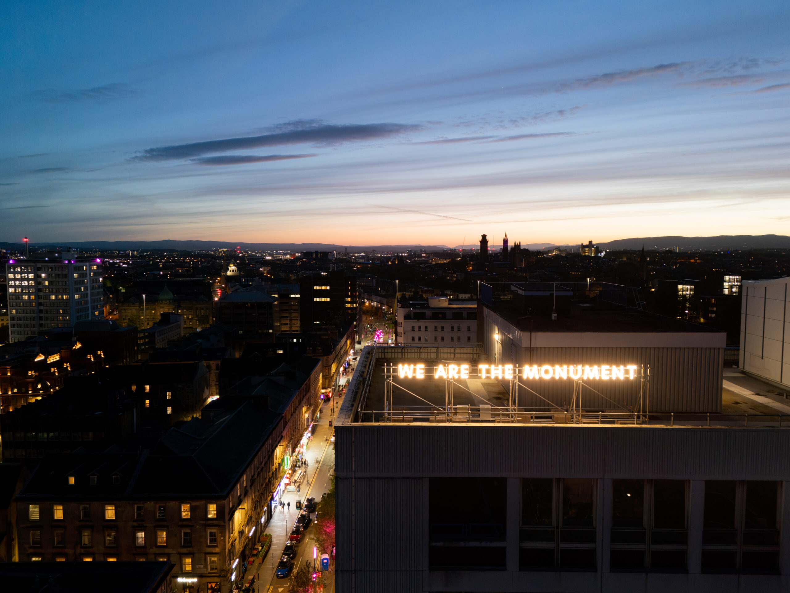 The artwork reading 'We are the monument' sits on the roof of Glasgow Dental Hospital and is illuminated against a night scene of Glasgow