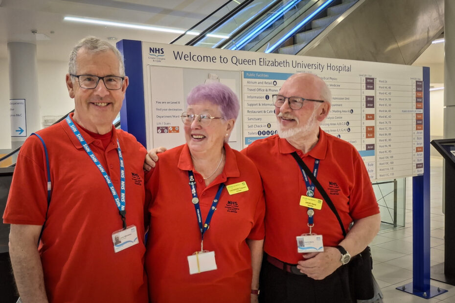 Three volunteers in the famous red T-shirts, smiling in front of an information sign at the Queen Elizabeth University Hospital