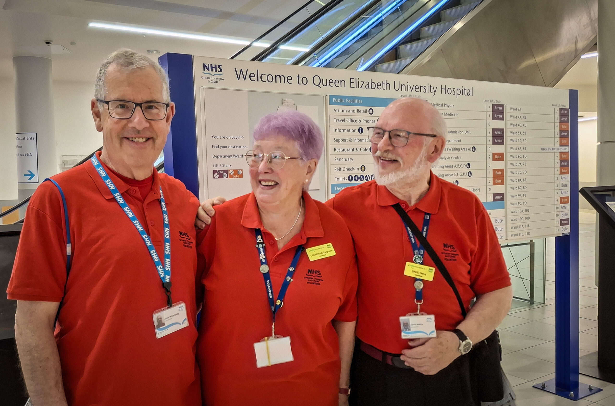 Three volunteers in the famous red T-shirts, smiling in front of an information sign at the Queen Elizabeth University Hospital