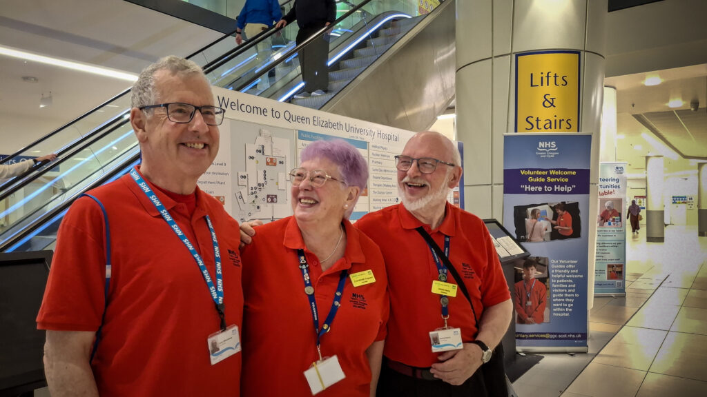 Three volunteers in the famous red T-shirts, smiling in front of an information sign at the Queen Elizabeth University Hospital