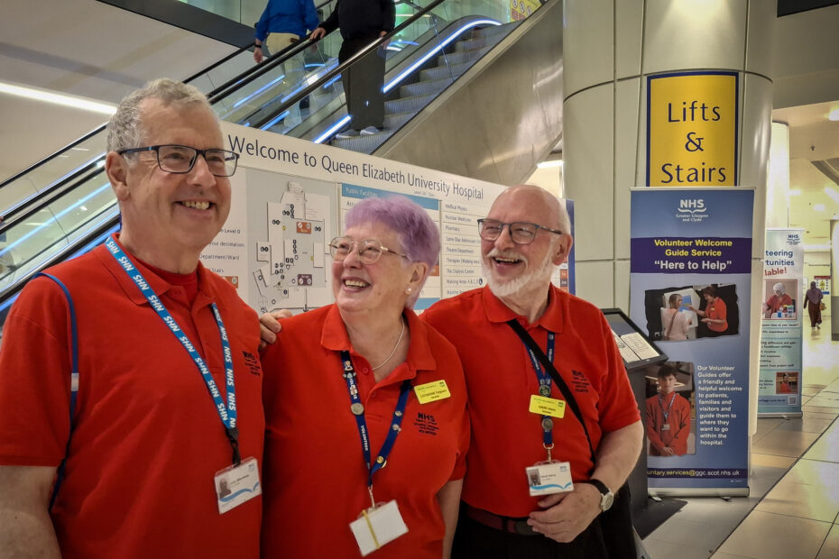 Three volunteers in the famous red T-shirts, smiling in front of an information sign at the Queen Elizabeth University Hospital