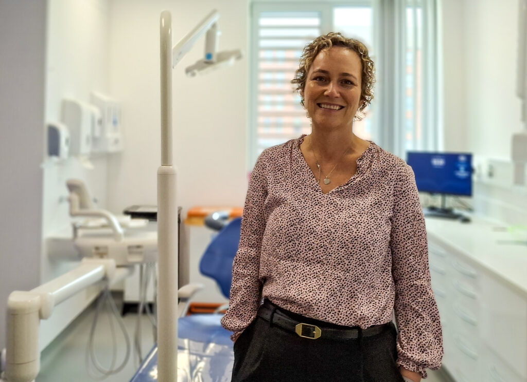 Kate Wiseman stands smiling in a treatment room, with the stand of a dental light next to her and a dental chair behind her.