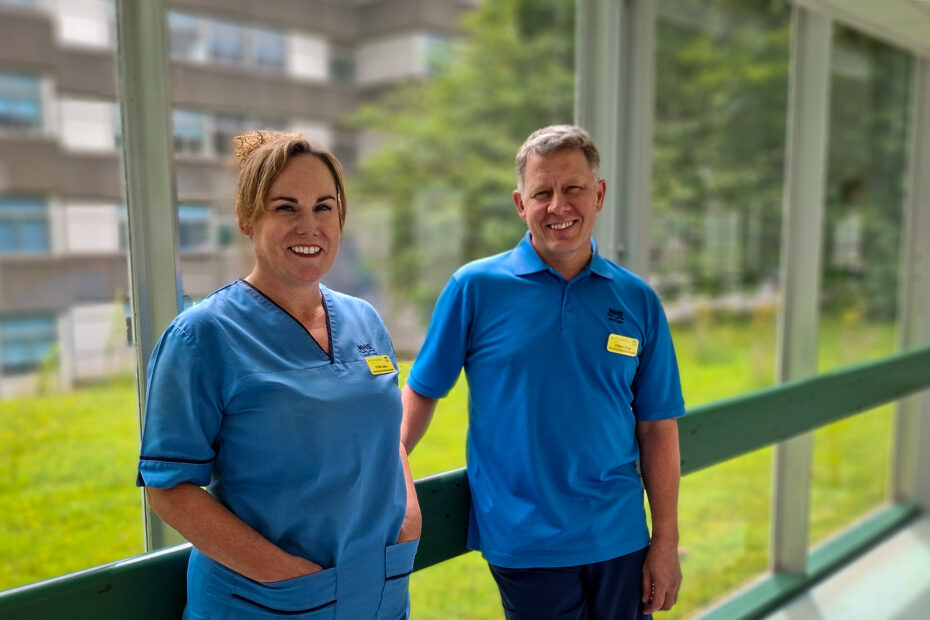 NHSGGC staff members Fiona Oakey and Joe Poole, both in blue uniforms, standing smiling in front of a large floor-to-ceiling wall of windows, with a view of hospital grounds outside the window.