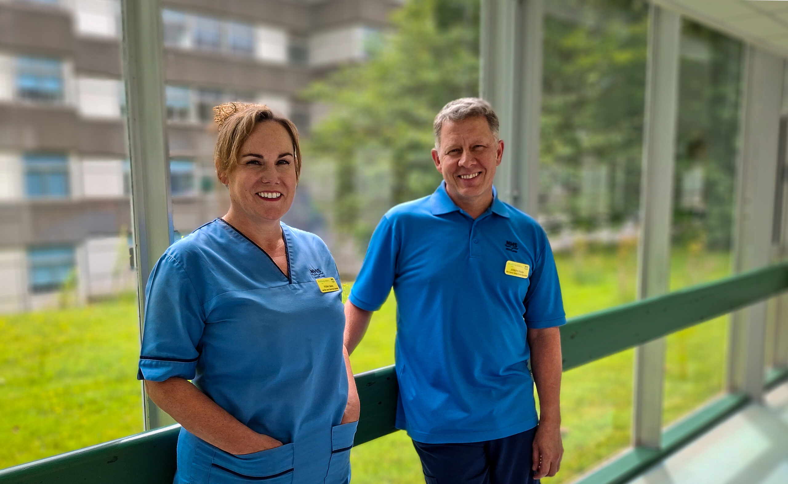 NHSGGC staff members Fiona Oakey and Joe Poole, both in blue uniforms, standing smiling in front of a large floor-to-ceiling wall of windows, with a view of hospital grounds outside the window.