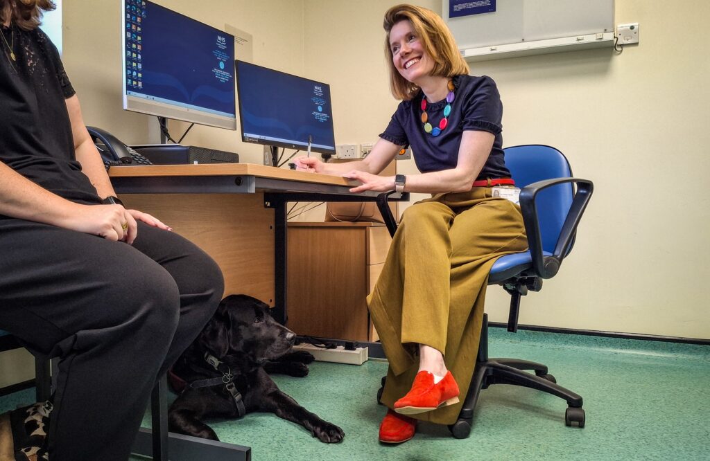 Dr Helen Grote sitting in a chair in front of a desk in a clinic room, during a consultation with a patient, partly visible on the left. Cooper the dog is lying under the desk.