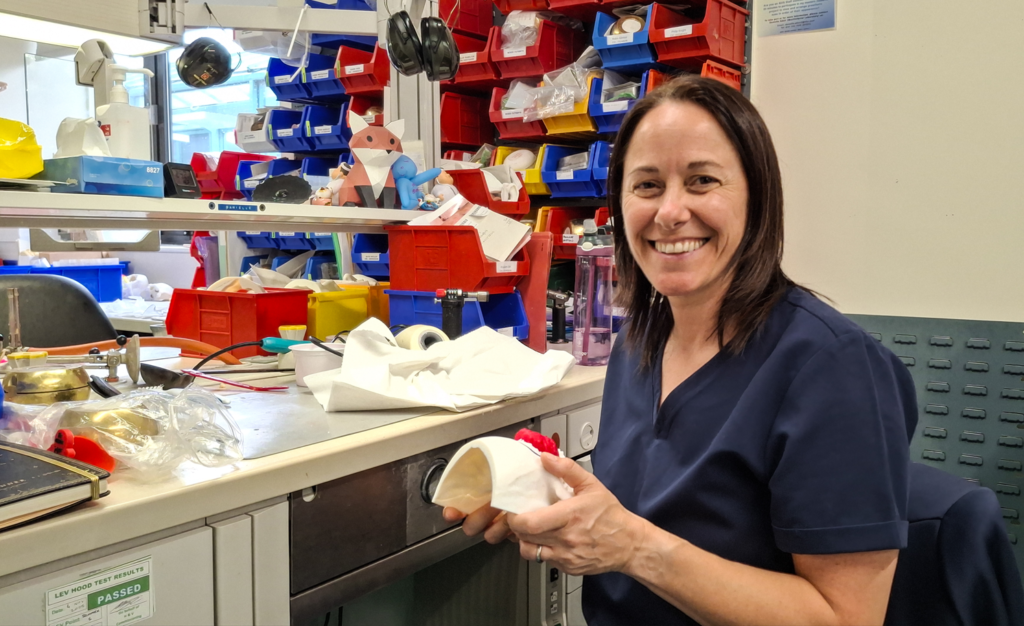 Principal Maxillofacial Prosthetist Elaine Baird in dark blue scrubs, holding a 3D printed model, sitting in front of a desk on the prosthetics lab.