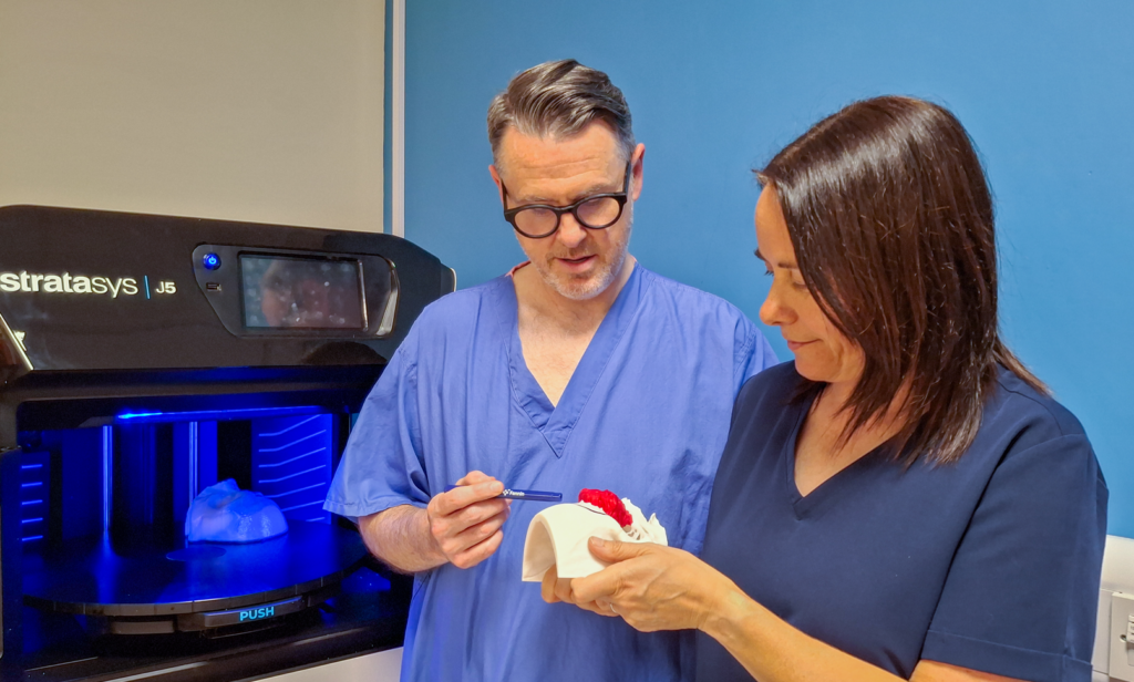 Elaine Baird and Craig Wales, both in blue scrubs, with a white facial model. In the background is the 3D printer
