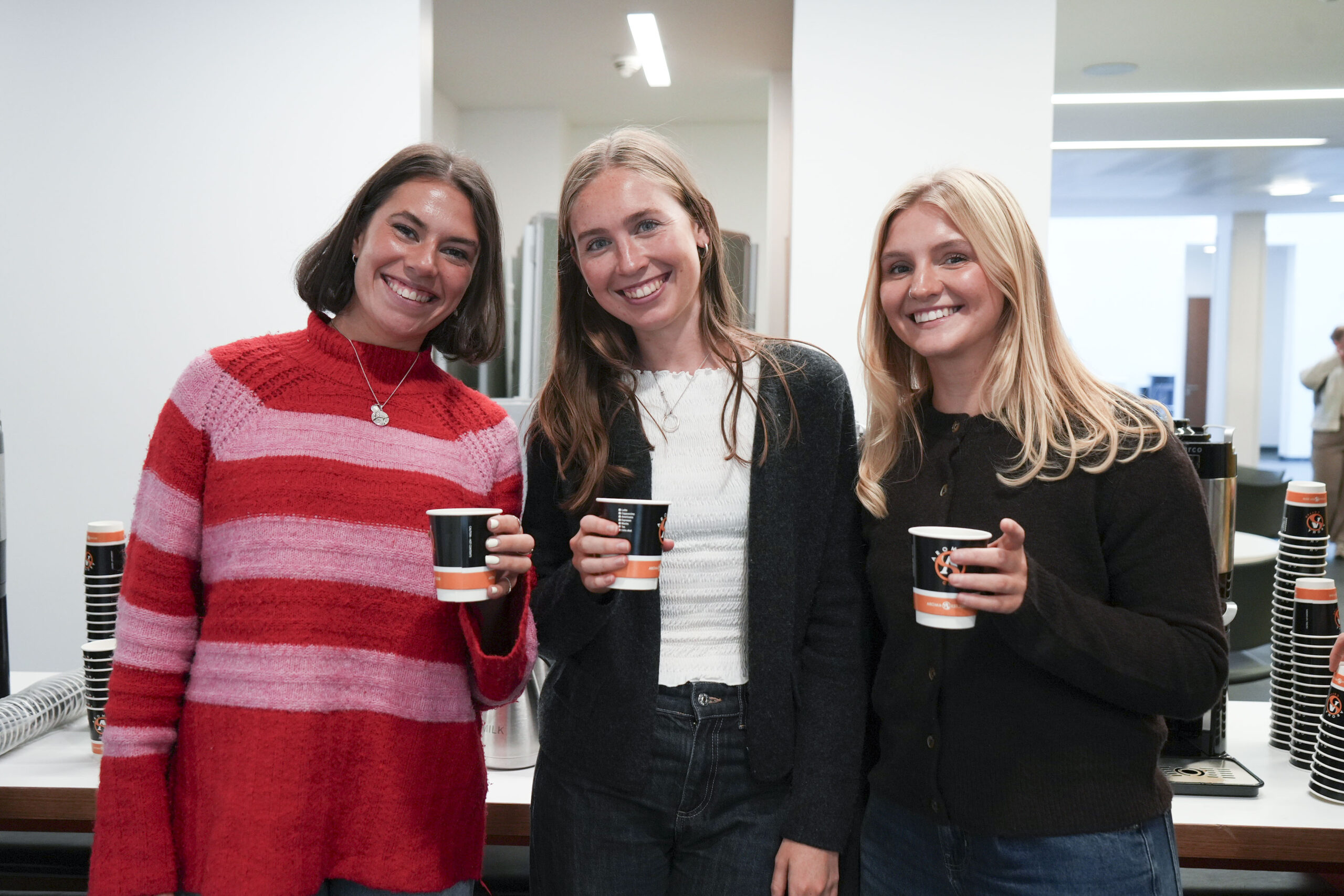 Three of the FY1s, each holding a coffee, smile and pose for the camera.