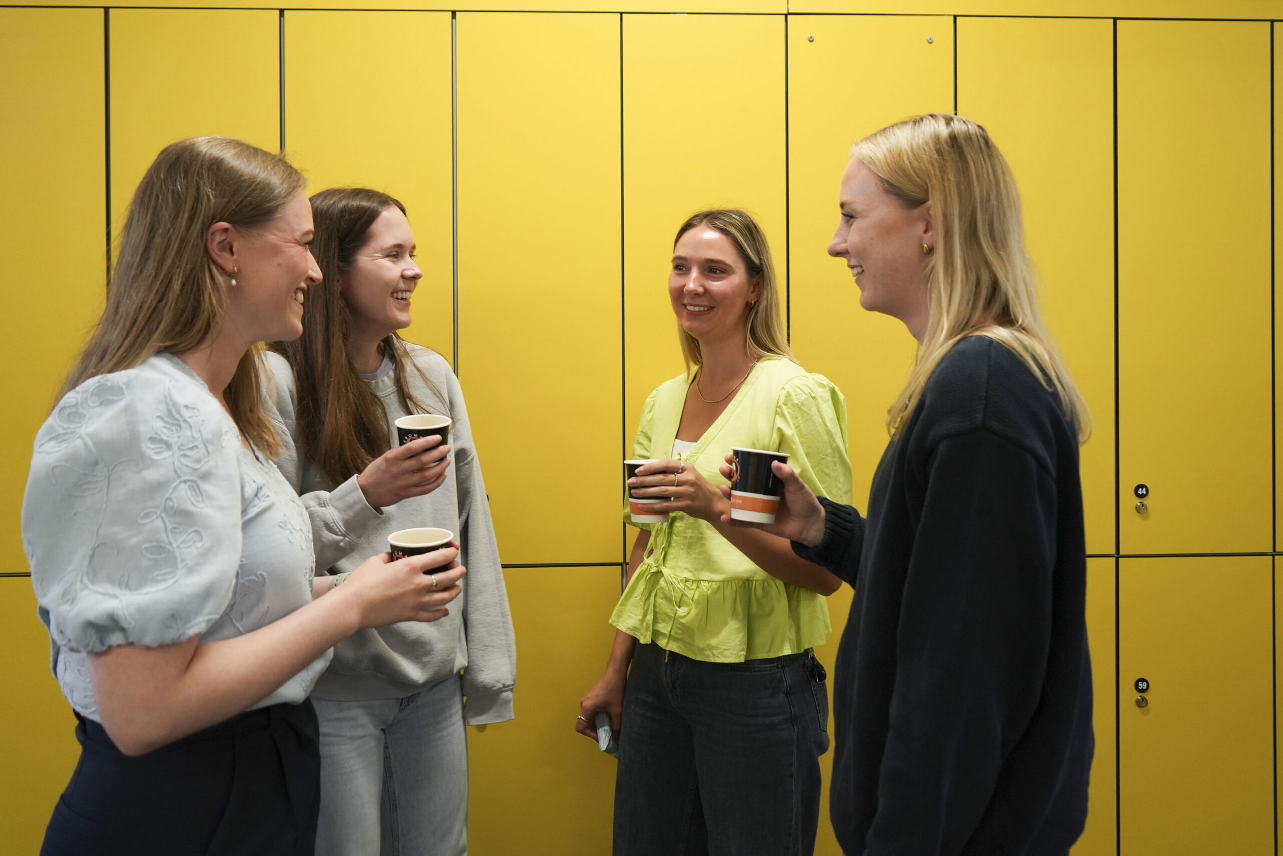 Four new doctors, each holding a coffee, chat in front of a wall of yellow lockers.