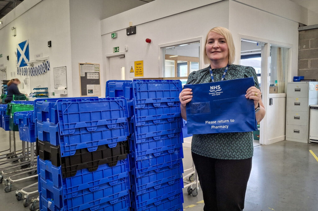Staff member Michelle Stevenson holds a blue medicine wallet while standing next to a pile of blue medicine delivery boxes. The picture was taken in the delivery hall of the Centralised Pharmacy Distribution Centre