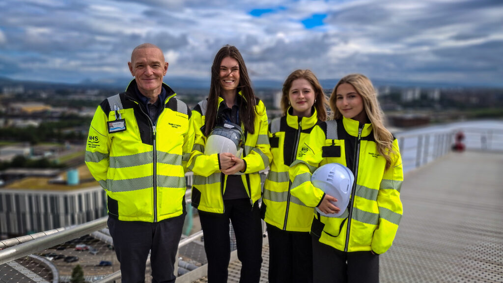 Errin, Amy and Ellie with Danny McInally (left). All are wearing yellow hi-vis jackets and Amy and Ellie are carrying hard hats. North-west Glasgow is in the background.