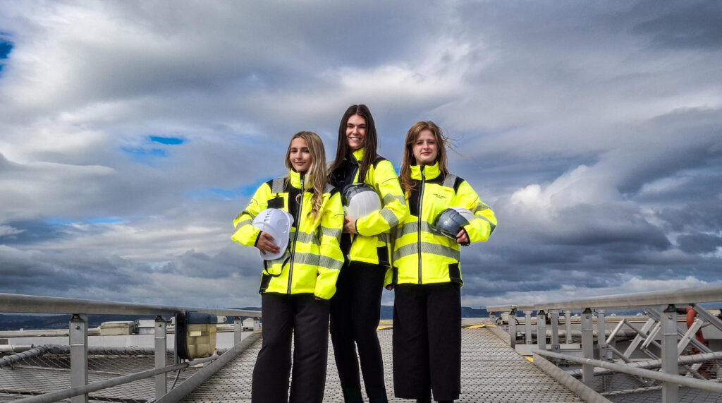 Ellie, Amy and Errin, all wearing yellow hi-vis jackets and are carrying hard hats, stand together against a background of a spectacular cloudy sky.