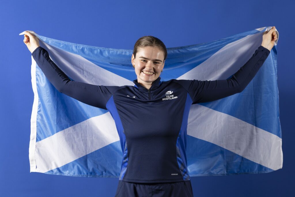 Rachel Conway in a blue Team Scotland tracksuit, stands smiling while holding a saltire behind her