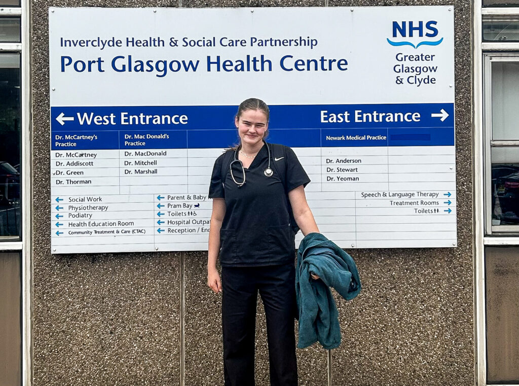 Rachel Conway, in black medical student's uniform, stands smiling in front of a sign for Post Glasgow Health Centre
