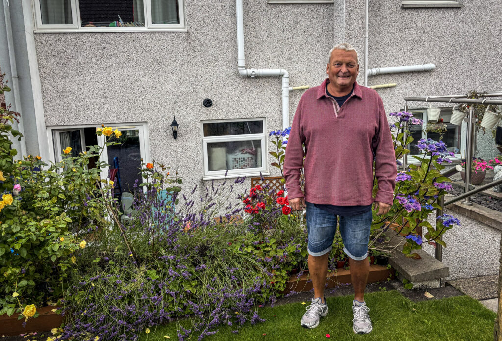 Robert Ormiston standing in his garden, with a flower bed behind and to the left of him and the back wall of his house behind that.