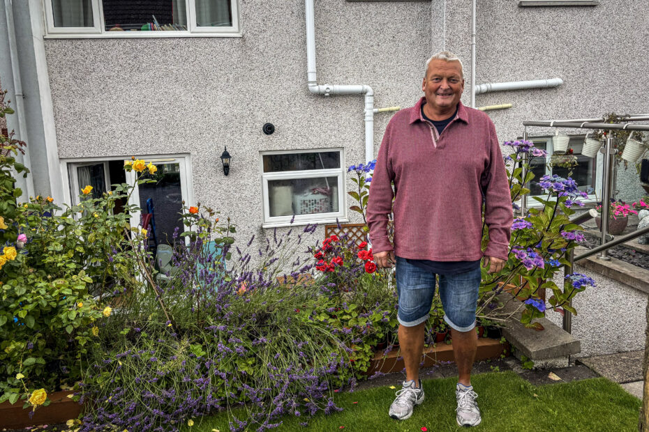 Robert Ormiston standing in his garden, with a flower bed behind and to the left of him and the back wall of his house behind that.
