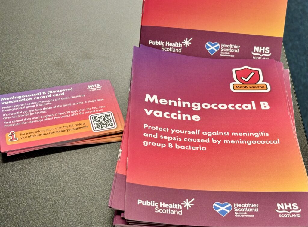 On the right is a pile of red-brown leaflets giving information on the Meningococcal B vaccine. To the left are small business cards with the same information
