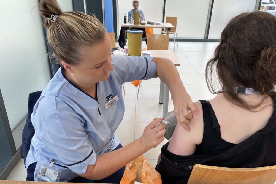 NHSGGC vaccinator in blue uniform administers the flu vaccine to a young woman at a vaccination clinic