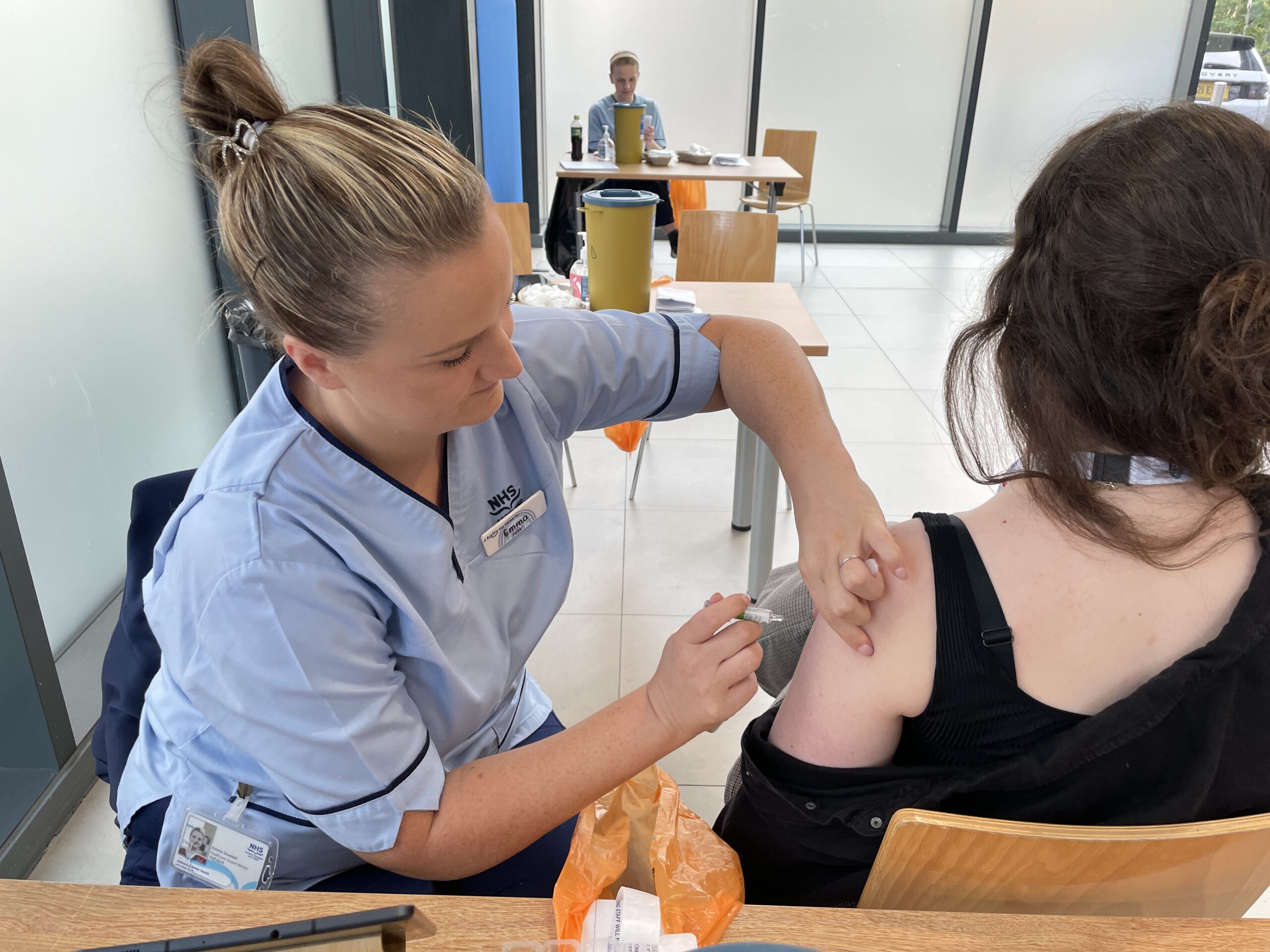 NHSGGC vaccinator in blue uniform administers the flu vaccine to a young woman at a vaccination clinic