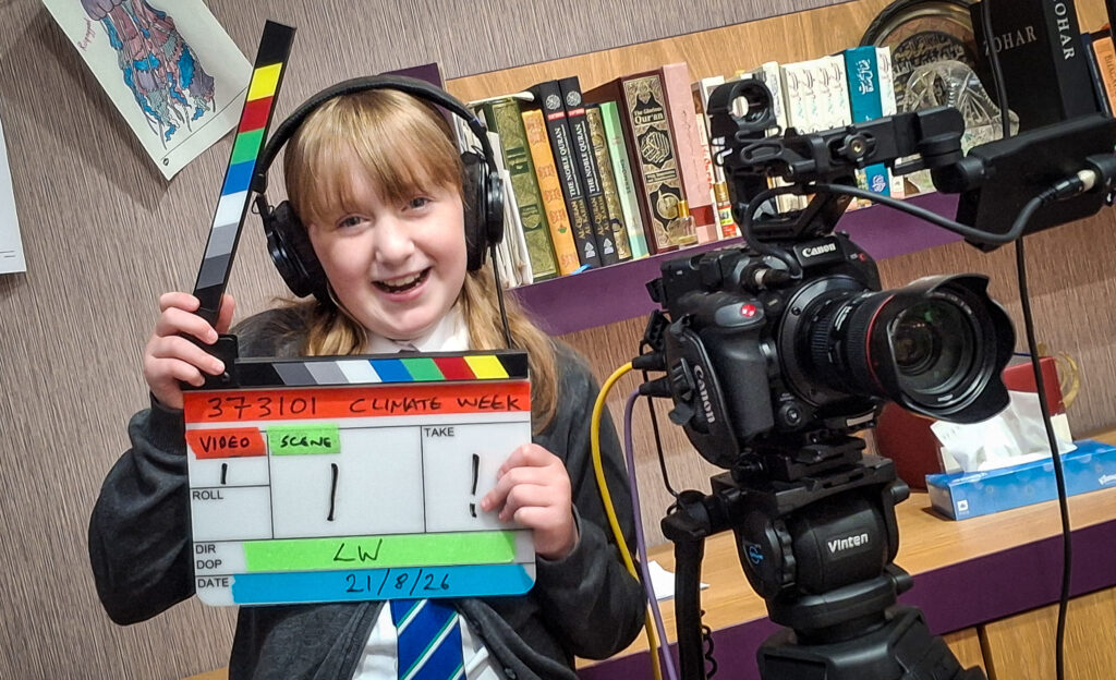 Flora Shelton, wearing headphones, smiles as he holds a traditional clapper board, while standing next to a video camera.