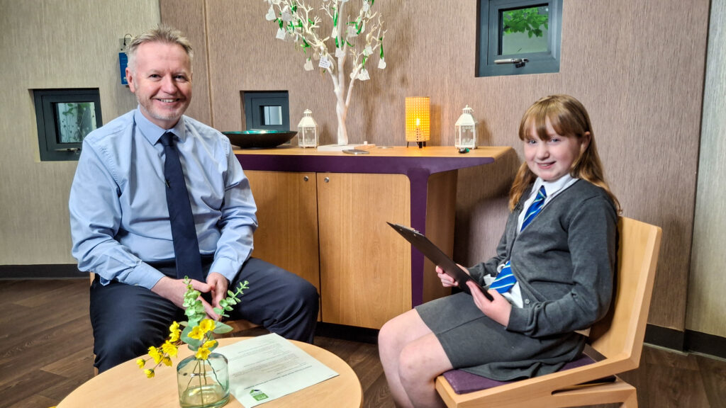 Michael Breen, left, wearing a blue shirt and tie, sits opposite Flora Shelton, with a low coffee table between them. Flora is holding a clipboard with questions she is about to ask Mr Breen.