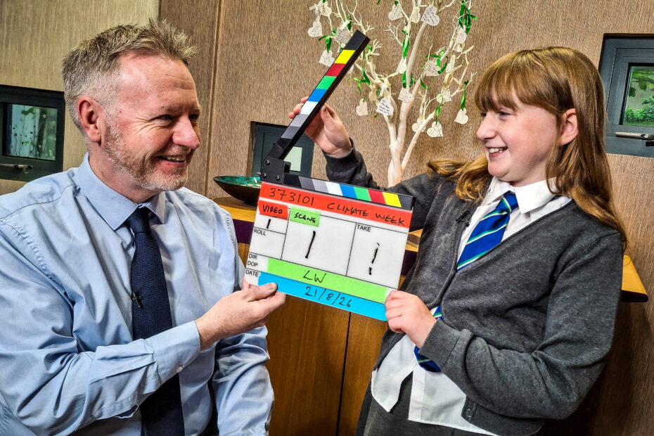 Michael Breen and Flora Shelton, both smiling and holding a clapper board, look at each other and smile ahead of Flora's interview.