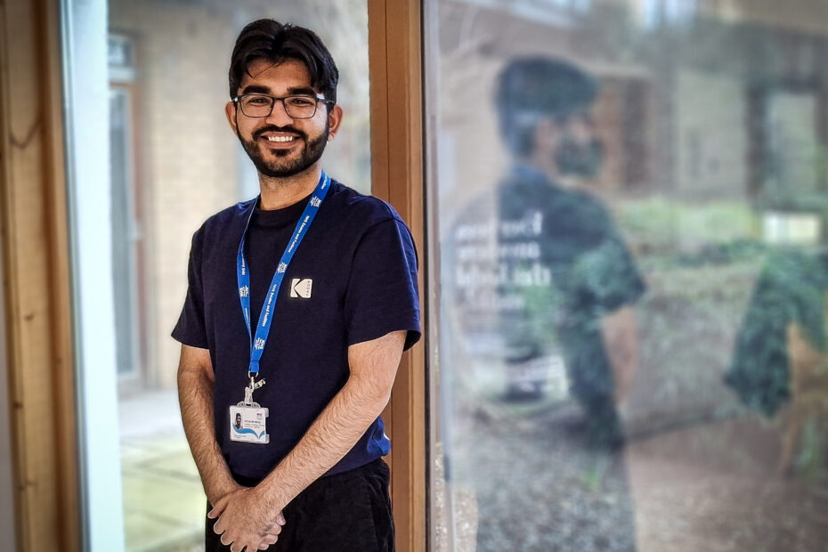 A smiling young man in a dark blue T-shirt with a lanyard around his neck stands in front of a large window with a garden outside.