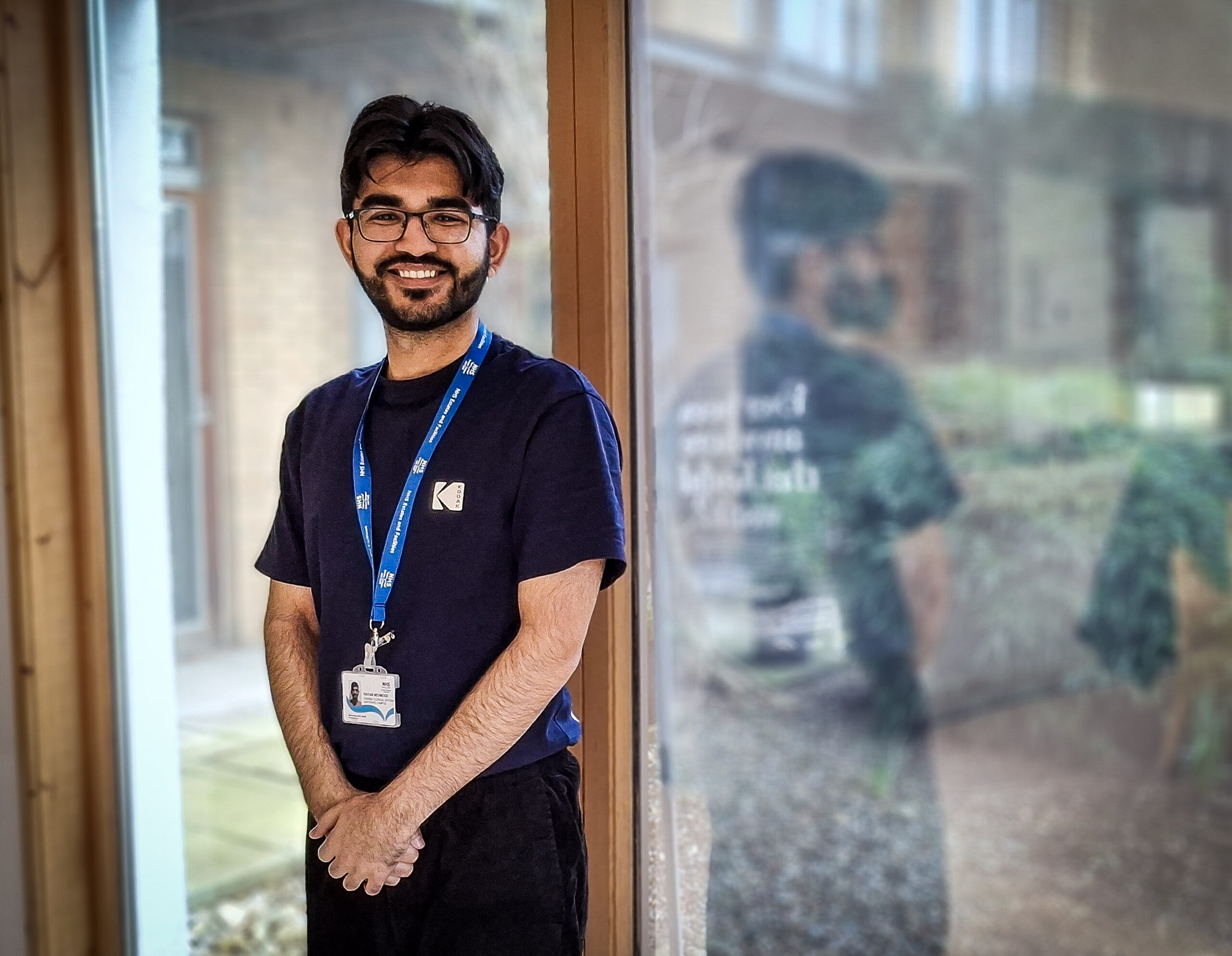A smiling young man in a dark blue T-shirt with a lanyard around his neck stands in front of a large window with a garden outside.
