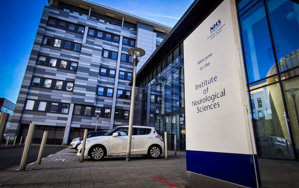 A general view of the Institute of Neurological Sciences, with a parked car in the bottom centre of the picture and a sign for the Institute in the foreground at the right.
