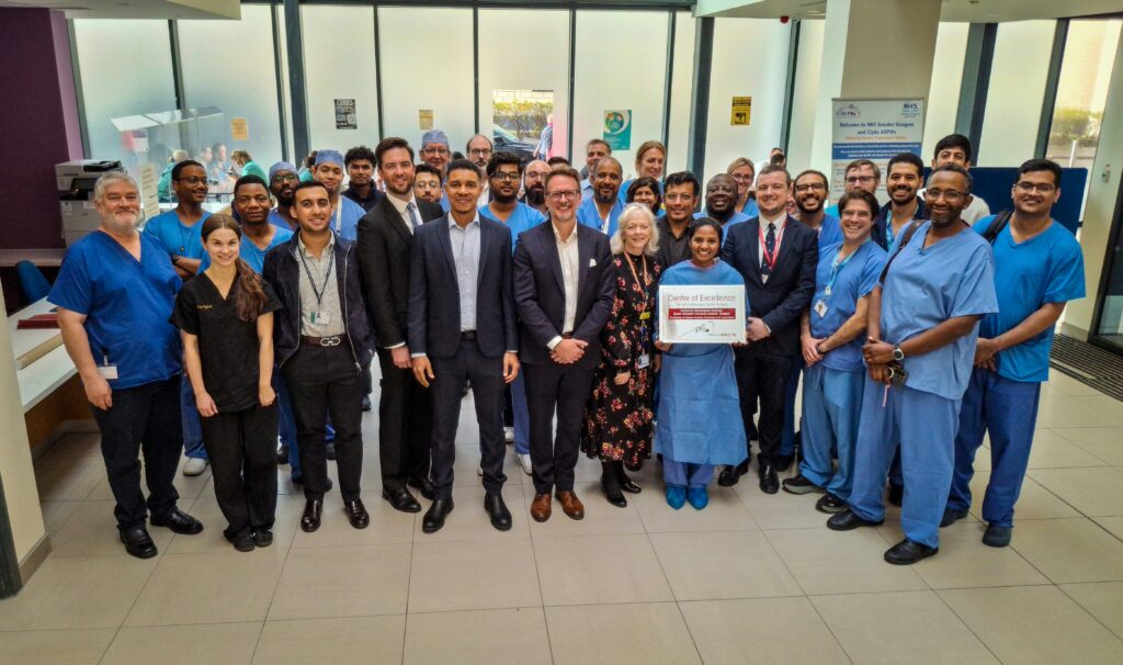 Members of staff, some in surgical uniform, gather in the atrium of the Institute of Neurological Sciences with the Centre of Excellence Award.