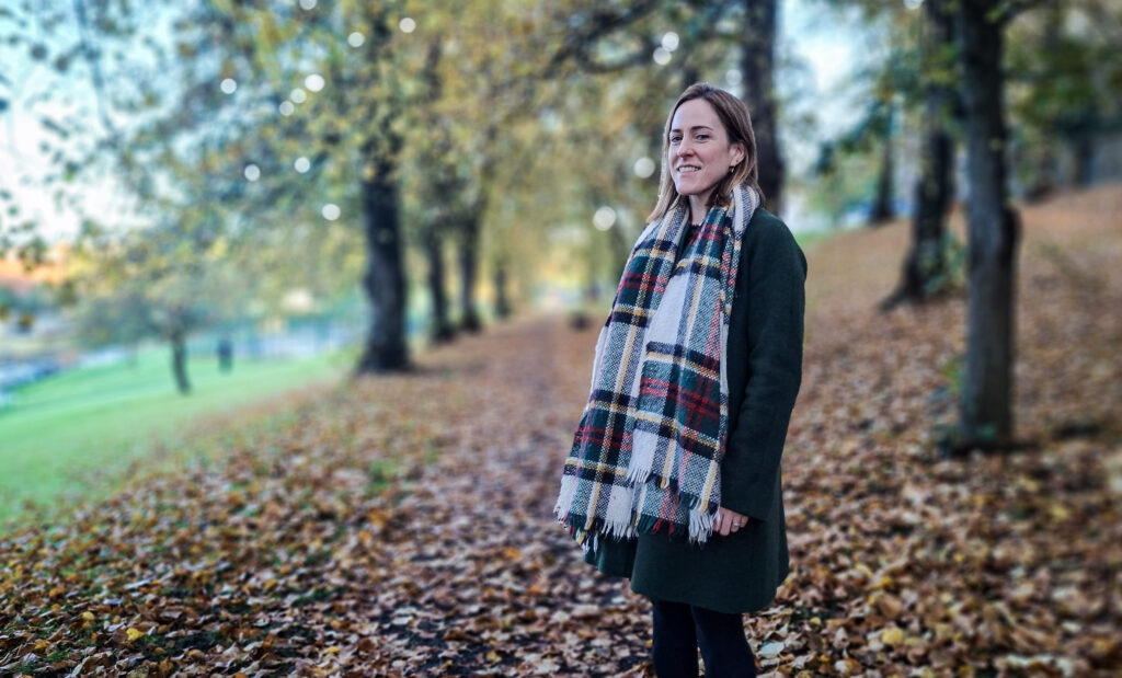 NHSGGC Head of Sustainability Keira McLuskey, in a coat and large tartan scarf, stands on a tree-lined path strewn with fallen leaves