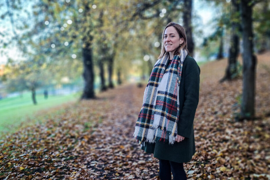 NHSGGC Head of Sustainability Keira McLuskey, in a coat and large tartan scarf, stands on a tree-lined path strewn with fallen leaves